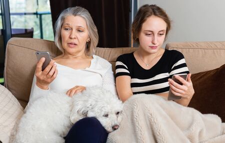 Young woman with elderly mother sitting with smartphones in home interiorの写真素材