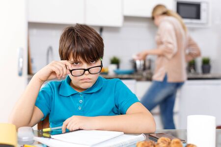 Portrait of boy studying at kitchen table and doing homework while his mother cookingの写真素材