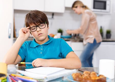 School age boy doing homework at kitchen while his mother preparing lunch at backgroundの写真素材