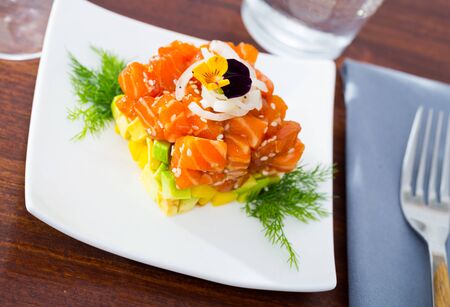 Fish tartare with raw salmon and diced avocado in shape of colorful cube decorated with pansy on white plateの写真素材
