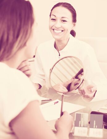 Young cheerful woman looking at mirror during consultation with cosmetologist in aesthetic medicine centeの写真素材