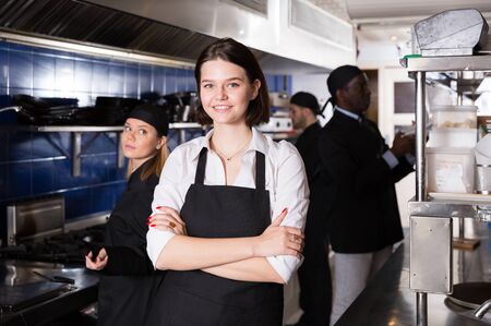Confident young waitress standing with arms crossed in modern kitchen with working staff of restaurantの写真素材