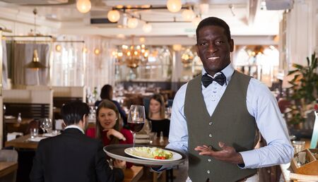 Glad waiter who is standing with order in hall of luxurious restaurantの写真素材