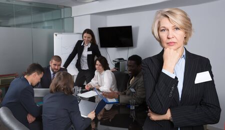 Upset mature businesswoman standing in meeting room on background with coworkersの写真素材
