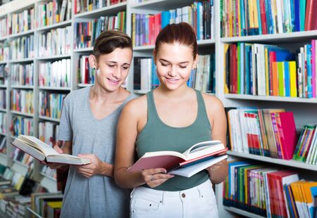portrait of smiling russian teenage boy and girl customers looking at open book standing among bookshelvesの写真素材