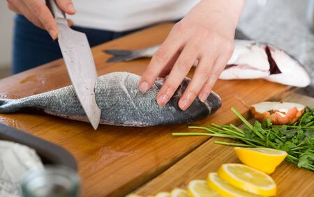 Woman cutting raw dorado fish on wooden board at kitchenの写真素材