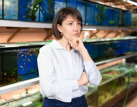 Young female aquarist standing near fish tanks in pet shop, thinking about purchase of tropical fish for home aquariumの写真素材