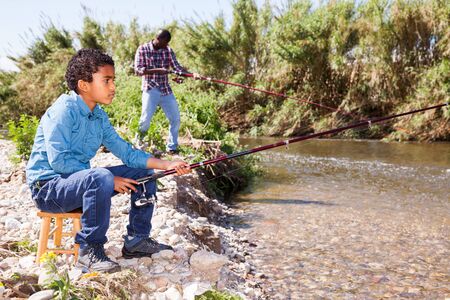 Positive African man and little boy standing near river and fishing with rodsの写真素材