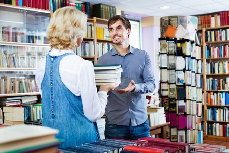 Young smiling man taking chosen book from seller in book shopの写真素材