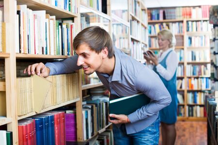 young  man in blue shirt standing among bookshelves in public library and searching for bookの写真素材