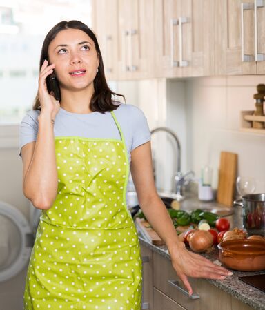 Portrait of young girl housewife in apron talking by phone at home kitchenの写真素材