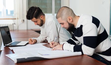 Two male university students sitting at desk preparing for exams together in classroomの写真素材