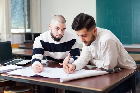 Two male university students sitting at desk preparing for exams together in classroomの写真素材