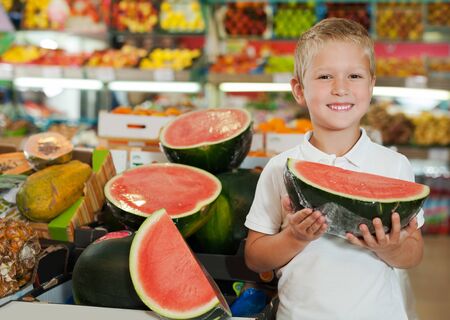 Portrait of cute cheerful kid holding piece of watermelon at fruit section of storeの写真素材
