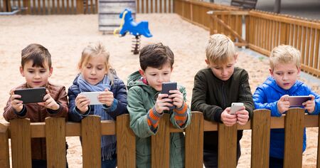 Group of preteen children standing near wooden fence on playground, addicted in their phonesの写真素材