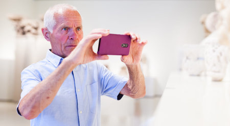 Mature male visitor using phone in sculptures exposition at museum