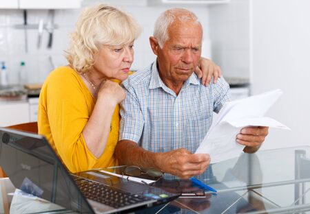 Worried mature couple with documents near laptop in home interior