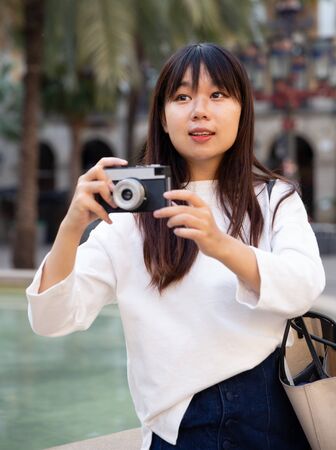 Young chinese woman traveler strolling with luggage around city, making photo of sightsの写真素材