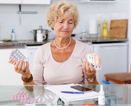 Mature woman holding blisters with pills and money in hands sitting at kitchen tableの写真素材
