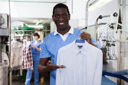 Smiling African-American man worker of laundry holding clean garments, demonstrating quality of dry cleaningの写真素材