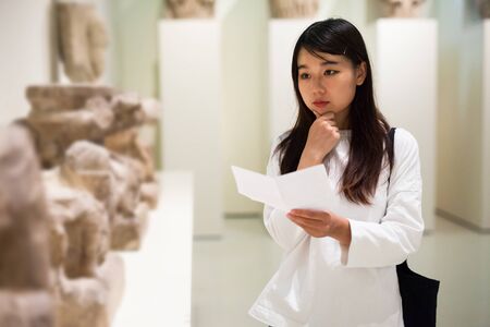 Young chinese woman visitor with  guide book looking at exhibition in museum of ancient sculptureの写真素材