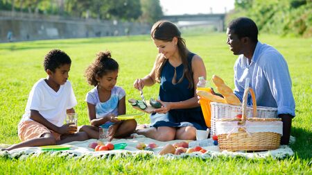 Smiling young parents with happy preteen daughter and son having fun together at picnic in summer city parkの写真素材