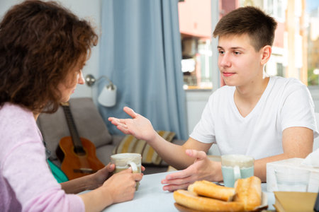 Cheerful teenage boy telling interesting story to his mother at home tableの写真素材