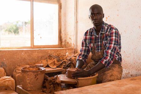 African young man focused on work on pottery wheel in workshop, creating new objectの写真素材