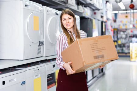 Satisfied pleasant smiling female housewife happy from buyings in home appliance store, standing with big boxの写真素材