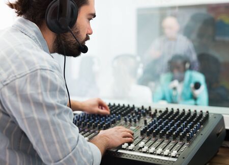 Focused bearded guy engaged in sound engineering, working at audio control panel in radio studioの写真素材