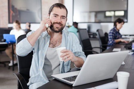 Smiling young Hispanic man talking on phone in busy modern open plan office