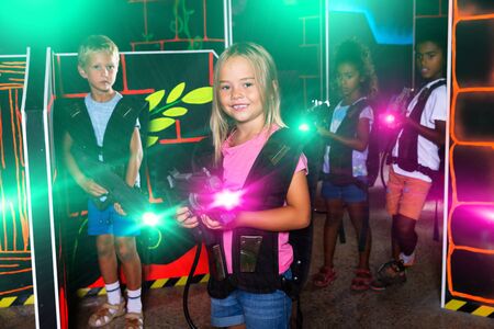 Cheerful little girl aiming laser gun at other players during lasertag game in dark roomの写真素材