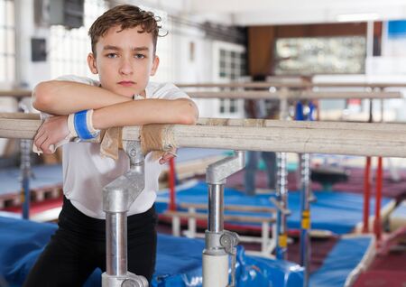 Unhappy tired teenage boy in sportswear standing near parallel bars at gymnastic hallの写真素材