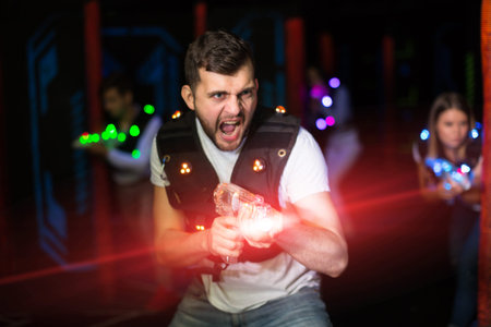 Emotional guy playing laser tag with friends on dark labyrinth in colorful beams of laser pistols