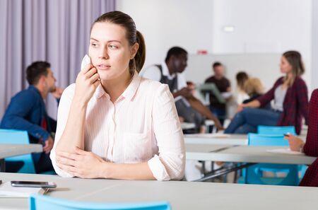 Upset girl sitting at table in auditorium during break on background with other studentsの写真素材