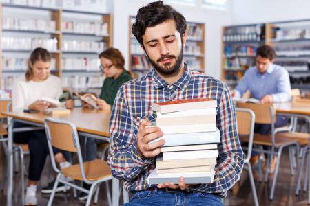Portrait of upset young man with stack of books in hands in public libraryの写真素材