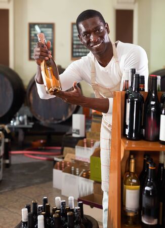Smiling diligent positive cheerful  salesman in apron proposing wine in bottles in winery shopの写真素材
