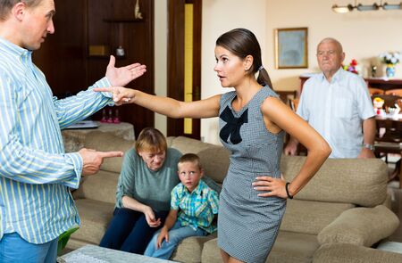 Portrait of young outraged woman emotionally talking to man on background with anxious senior parents and tween boyの写真素材