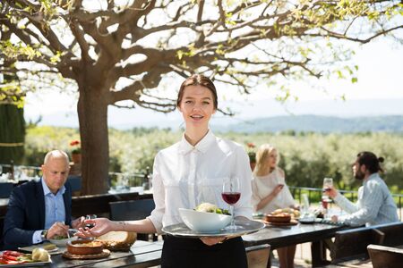 Hospitable young female owner of cosy country restaurant in open air meeting guests with serving tray in handsの写真素材