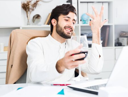 Positive young male manager with glass of wine having emotional online conversation at office deskの写真素材