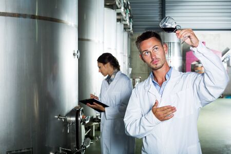 Portrait of cheerful young man in uniform looking at wine sample in glass on wine factoryの写真素材