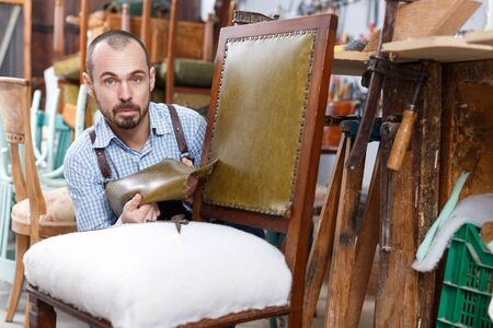 Professional male carpenter upholstering vintage armchair in repair shopの写真素材