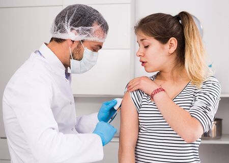 Smiling man doctor making injection of vaccine to teenager girl patient in hospitalの写真素材