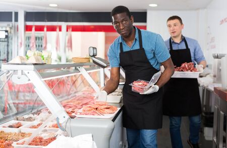 Two professional butchers arranging meat products in display case of butcher shopの写真素材