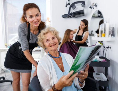 Young woman hairdresser in hair salon helping to senior woman choosing hairstyling in magazine
