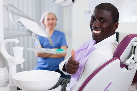 Happy african american man sitting in dental chair after teeth cure giving thumb upの写真素材
