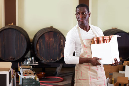 Diligent cheerful smiling afro seller working in wine store, holding box full of bottles with wine