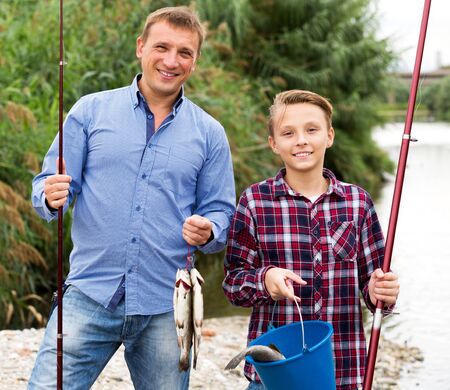 Portrait smiling man with teenager boy standing together and showing catch fish outdoorsの写真素材