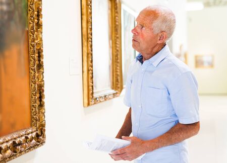 Intelligent mature tourist holding brochure with exhibition program near paintings in modern museumの写真素材