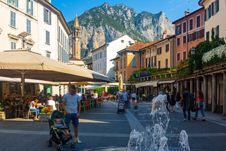 View of busy central street of Italian city of Lecco with gothic bell-tower of Minor Basilica of San Nicolo on background of San Martino mountain in sunny summer dayの写真素材